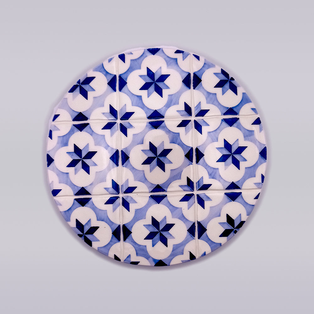 A round pattern featuring a series of blue and white geometric floral designs. The hand-made pattern is repetitive with an eight-pointed star shape in the center of each flower-like motif, set against a light grey background, perfect for adding charm to your kitchen decor as a Rossio Ceramic Trivet for Hot Pans by Tejo Shop.