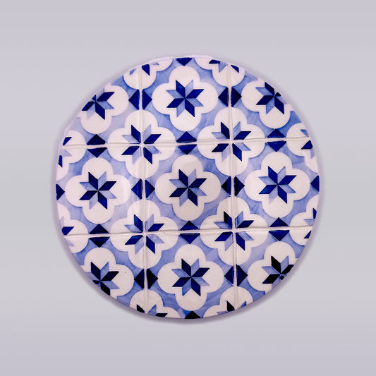 A round pattern featuring a series of blue and white geometric floral designs. The hand-made pattern is repetitive with an eight-pointed star shape in the center of each flower-like motif, set against a light grey background, perfect for adding charm to your kitchen decor as a Rossio Ceramic Trivet for Hot Pans by Tejo Shop.