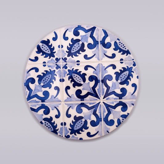 A hand-crafted Tejo Shop Sagres Ceramic Pad for Hot Pans showcases a circular decorative tile with a repeating pattern of blue floral and abstract designs on a white background, creating an intricate and symmetrical motif across the surface. This exquisite piece is meticulously hand-painted, adding a touch of artisanal elegance to any space.