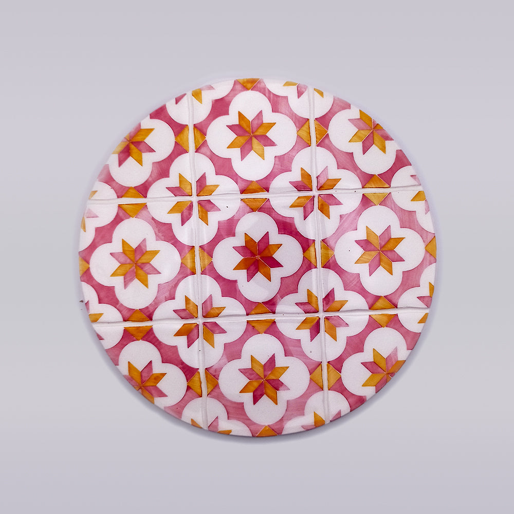 A hand-crafted round object featuring a colorful geometric pattern. The pattern includes star shapes in shades of pink, orange, and white on a light gray background. This Sines Ceramic Trivet Stand by Tejo Shop design repeats symmetrically across the entire surface, making it an elegant ceramic tile trivet.