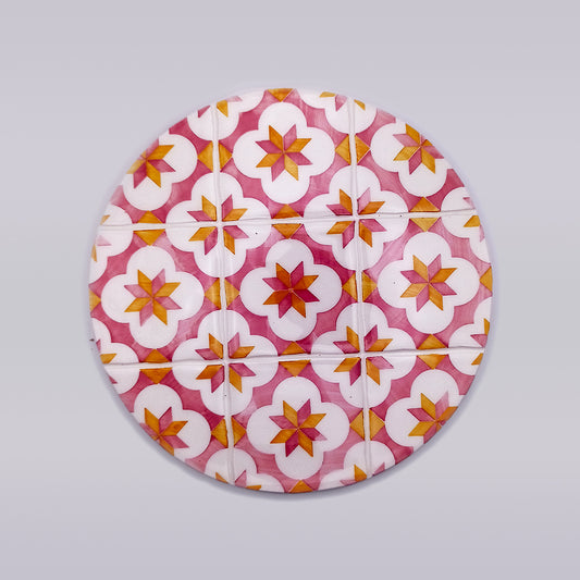A hand-crafted round object featuring a colorful geometric pattern. The pattern includes star shapes in shades of pink, orange, and white on a light gray background. This Sines Ceramic Trivet Stand by Tejo Shop design repeats symmetrically across the entire surface, making it an elegant ceramic tile trivet.
