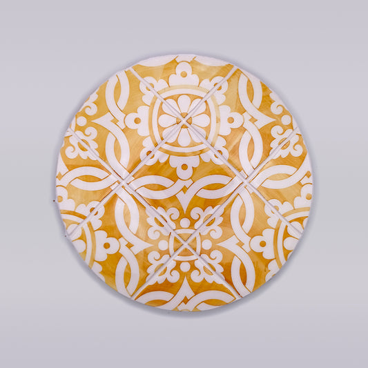 A round, ornate hand-painted trivet features an intricate white and yellow geometric pattern. The design includes symmetric floral and vine motifs set against a warm yellow background, creating an elegant and decorative appearance perfect for kitchen decor. The Sintra Ceramic Kitchen Trivet by Tejo Shop is displayed on a plain gray surface.