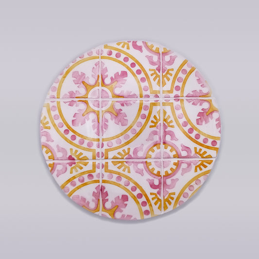 A round coaster with an intricate tile pattern featuring pink and yellow geometric and floral designs. The coaster, reminiscent of a Tavira Ceramic Tile Trivet from Tejo Shop, is divided into a grid of smaller square tiles, each continuing the overall motif. Hand-made with quality ceramic tiles, the background is plain light grey.