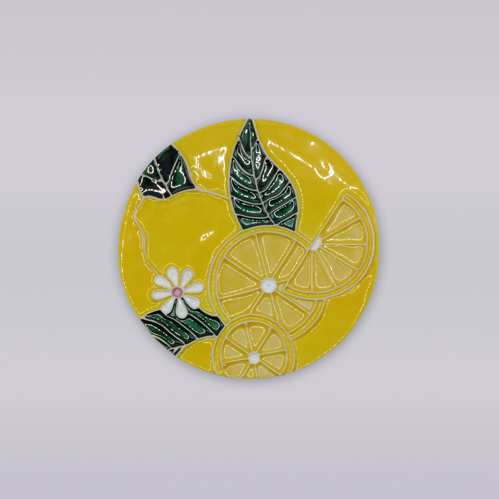 The Tejo Lemon Blossom Coaster – Ribatejo Edition by Miradouro Ceramics features sliced lemons, green leaves, and white flowers on a round yellow ceramic surface inspired by traditional Portuguese designs.