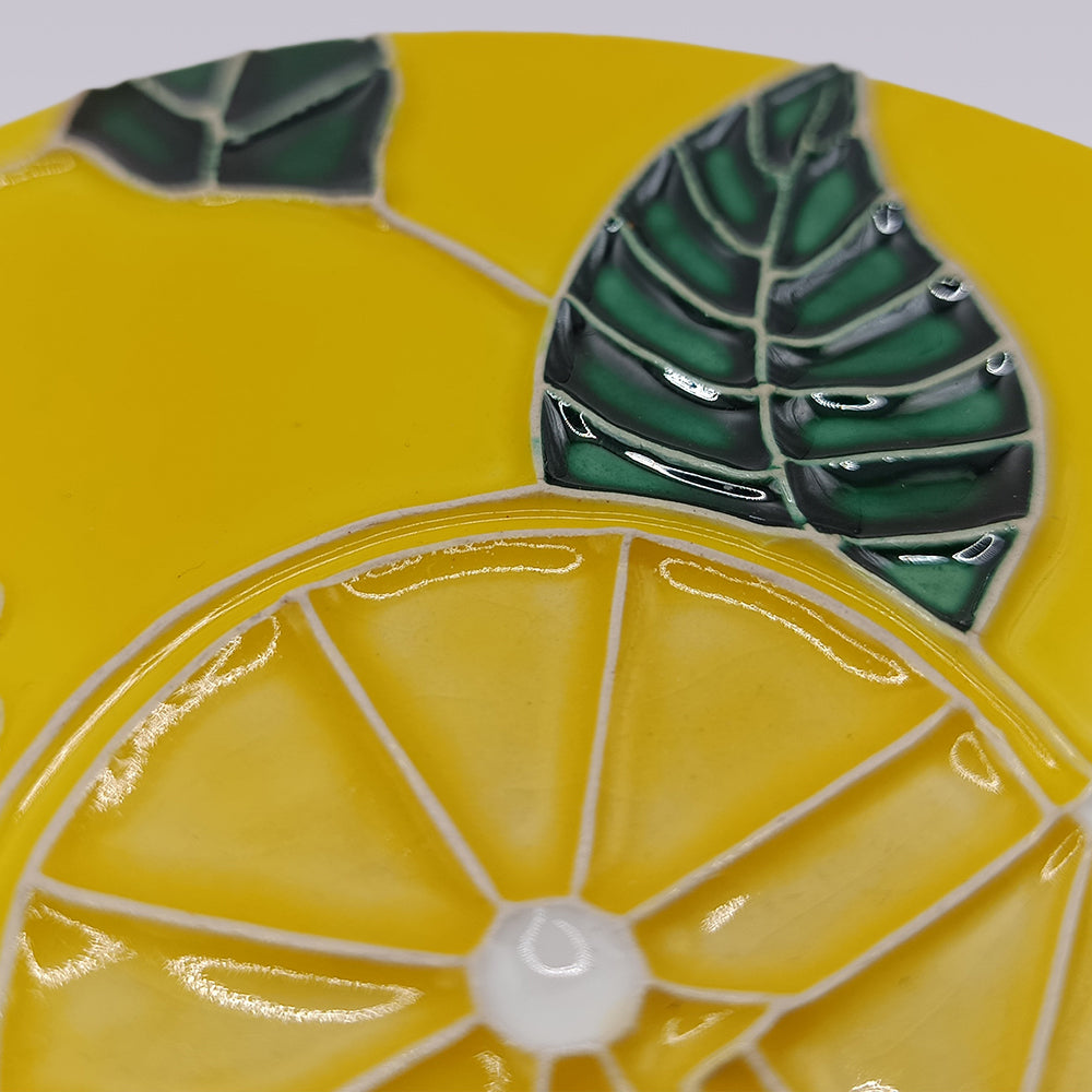 A close-up of the Tejo Lemon Blossom Coaster – Ribatejo Edition by Miradouro Ceramics, a Portuguese ceramic coaster with a raised lemon slice and two green leaves on a vibrant yellow background.