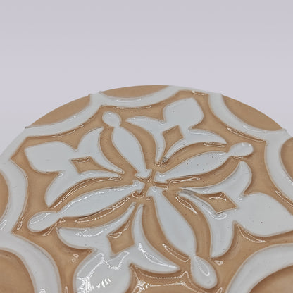 A close-up of the Terra Manuelina coaster by Miradouro Ceramics, handcrafted with a raised glossy white floral pattern on matte light brown, featuring symmetrical petals inspired by Portuguese ceramics.