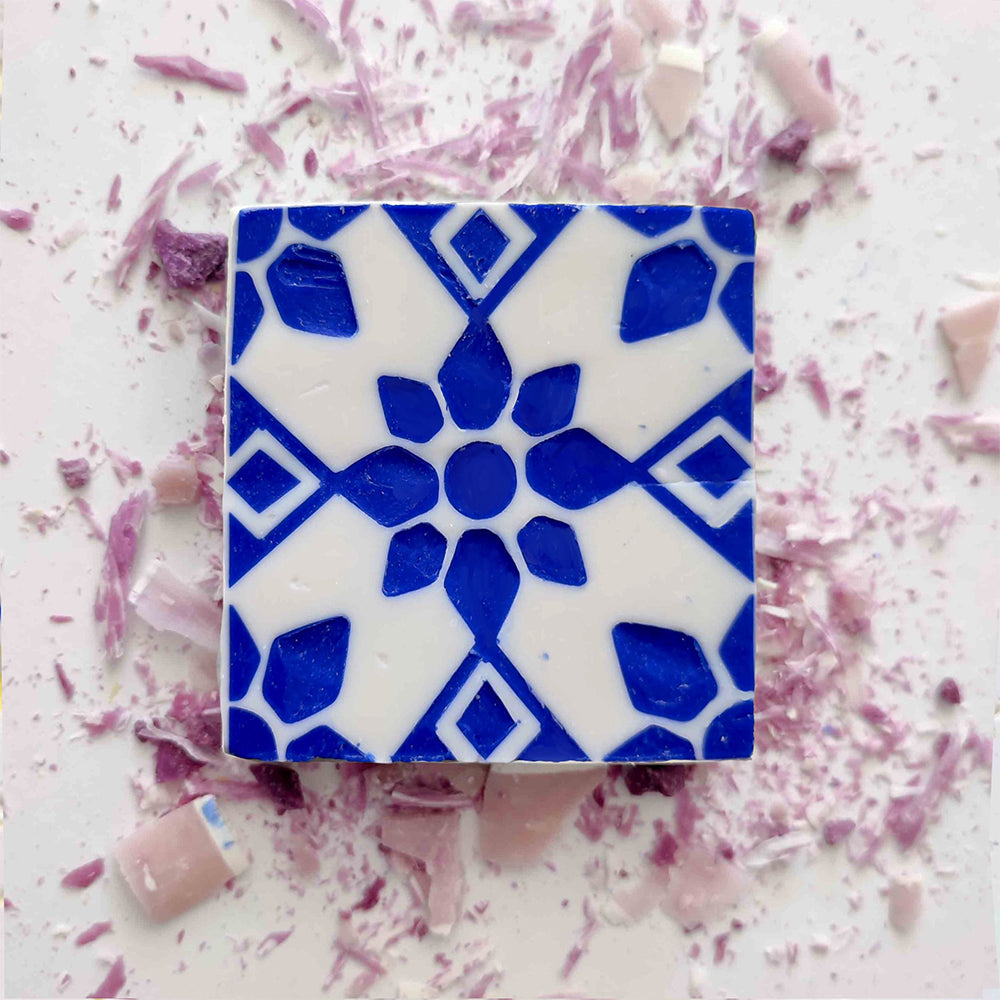 The Lisbão Violet Azulejo Ritual soap features a blue geometric floral design on a white square, set amid pink and purple tile fragments, capturing the artisan allure of traditional Portuguese tiles.