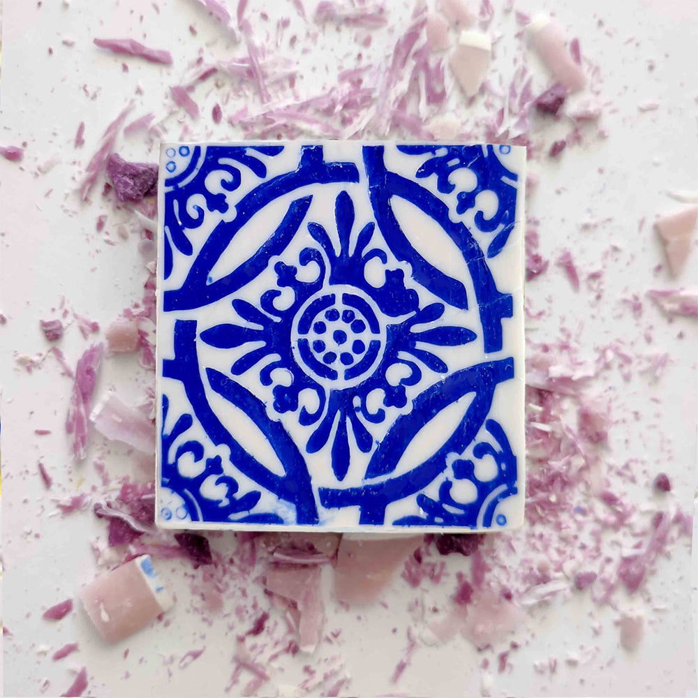 The Lisbão Violet Azulejo Ritual soap, a square handmade Portuguese tile design with an intricate blue floral pattern, rests on a white surface dotted with pink and purple shards.