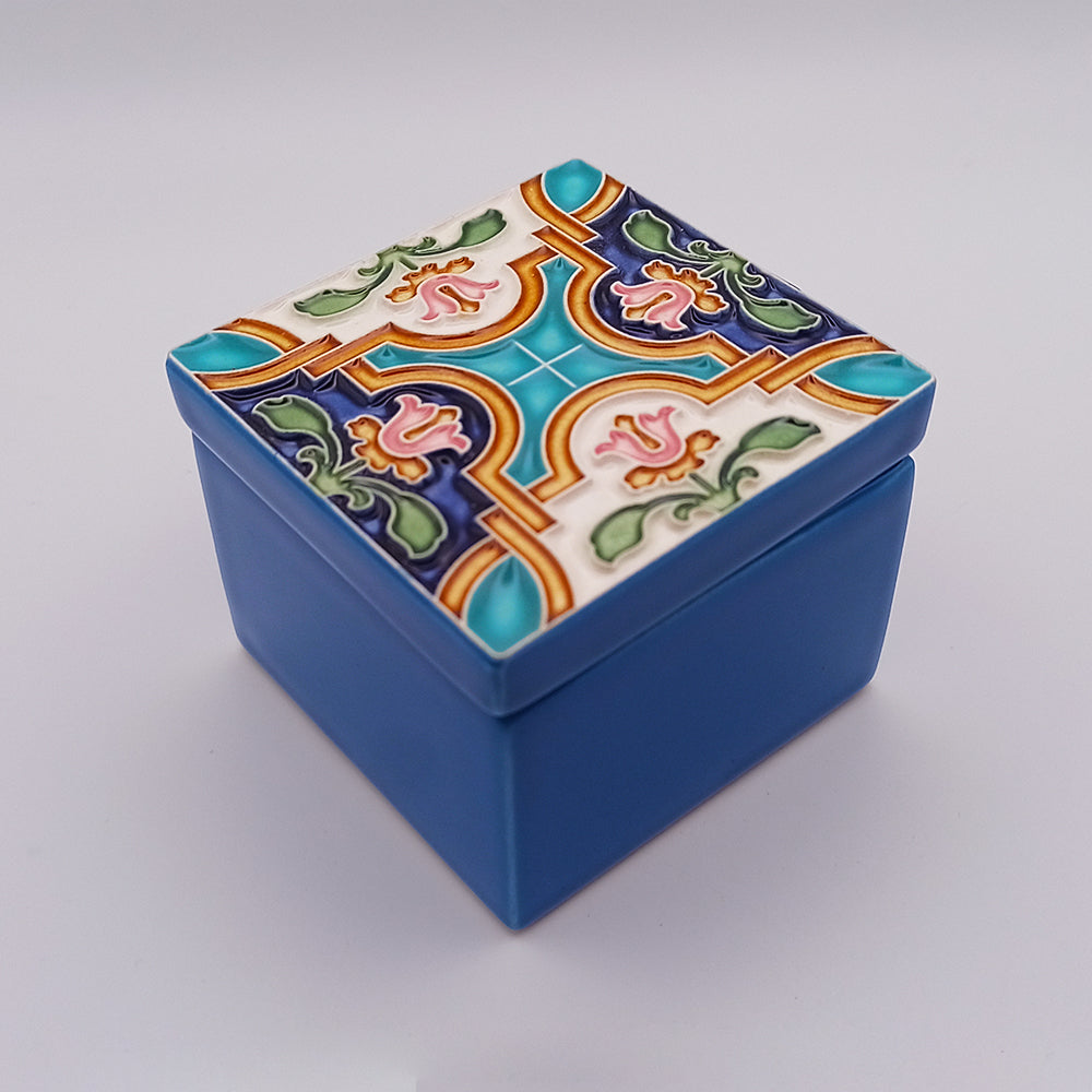 A small blue square Albufeira Ceramic Box from Tejo Shop with a decorative lid. The handcrafted lid boasts an intricate tile design with colorful floral and geometric patterns in blue, green, pink, and white. This unique addition rests elegantly on a light gray background.