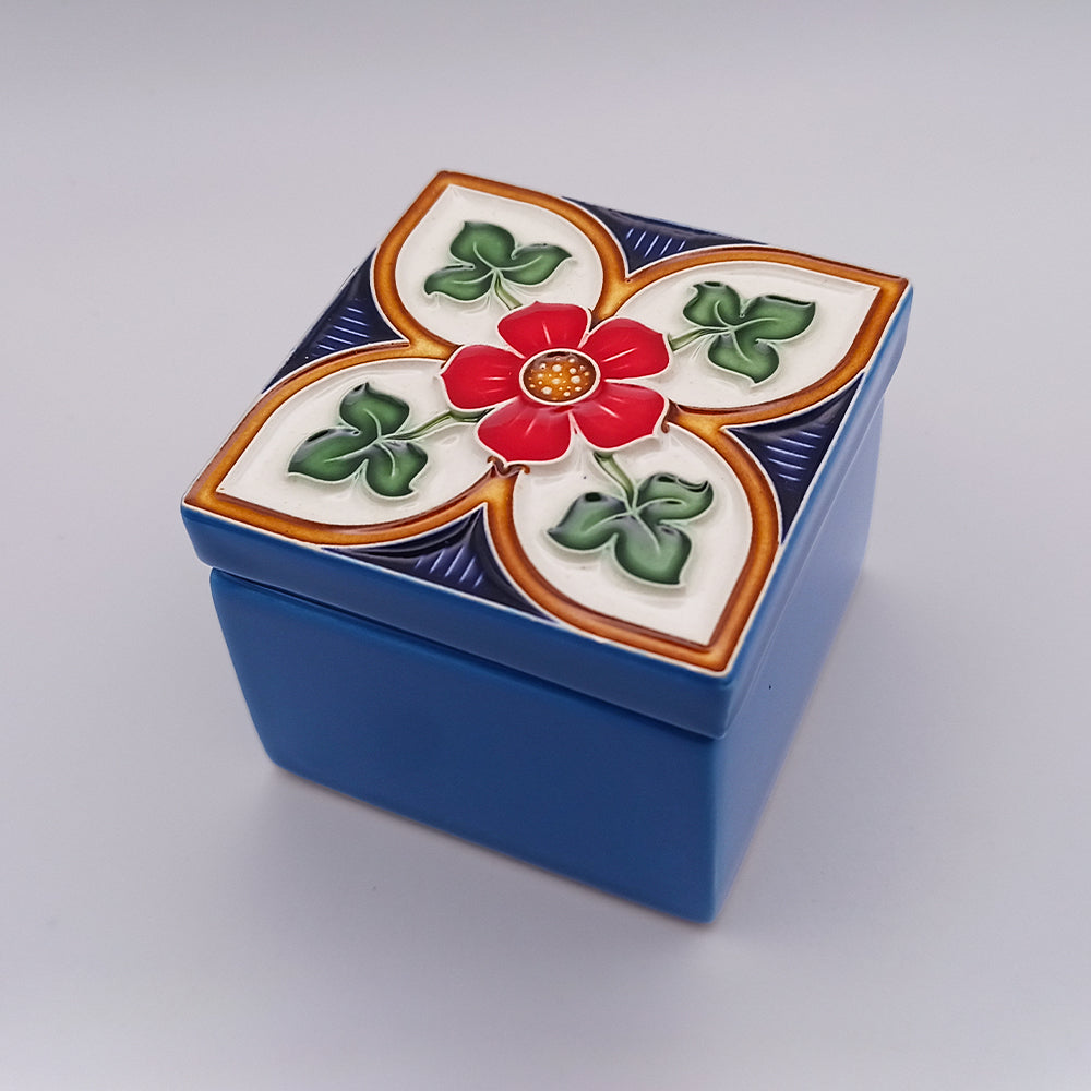 A small, square blue decorative box with a colorful lid. Handcrafted, the Alcobaça Ceramic Box by Tejo Shop features an ornate design with a central red flower surrounded by green leaves and white petal patterns, bordered by orange and dark blue accents. The background is a plain gray.