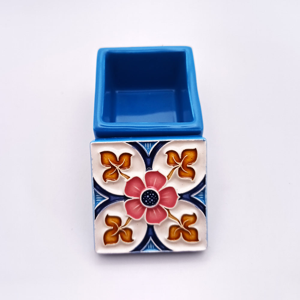 The Alfama Ceramic Box by Tejo Shop is a small, blue ceramic container perfect for home decor, adorned with an ornate, colorful floral design on one side. This handmade piece features a central pink flower surrounded by four smaller, golden-yellow flowers set against a white background with blue decorative accents.