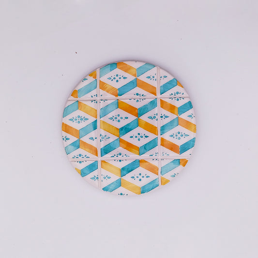 A hand-made, round ceramic tile with a geometric pattern in blue, turquoise, and yellow tones. The hand-painted design features alternating diamond shapes and small symmetrical flower-like motifs within some diamonds, creating a vibrant and decorative Arraiolos Ceramic Tile Cup Pad by Tejo Shop.