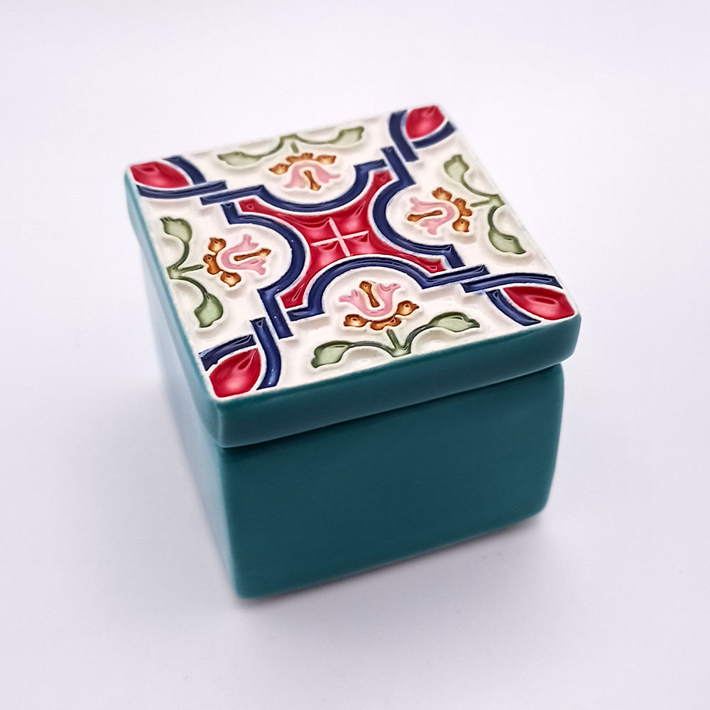 A small, square **Azores Islands Ceramic Box** with a teal base and a decorative, multicolored lid featuring an intricate symmetrical pattern in red, blue, green, and white. The exquisite lid design has a mixture of floral and geometric motifs, reminiscent of the artistic details found in Azores Islands ceramic art. Available at **Tejo Shop**.