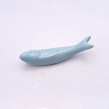 A small, light blue ceramic fish from Tejo Shop's collection rests on a plain white background. The Ceramic Sardine Fridge Magnet boasts a simple and streamlined design with minimal detailing to suggest scales and fins, embodying clean, modern lines that make it a charming addition to any kitchen décor.