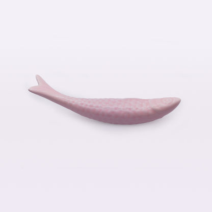 A small, pink ceramic object shaped like a fish, reminiscent of Portuguese Ceramic Sardines by Tejo Shop, lying flat on a white surface. It has a rounded head and tapering tail, with subtle, textured details to represent scales along its body.