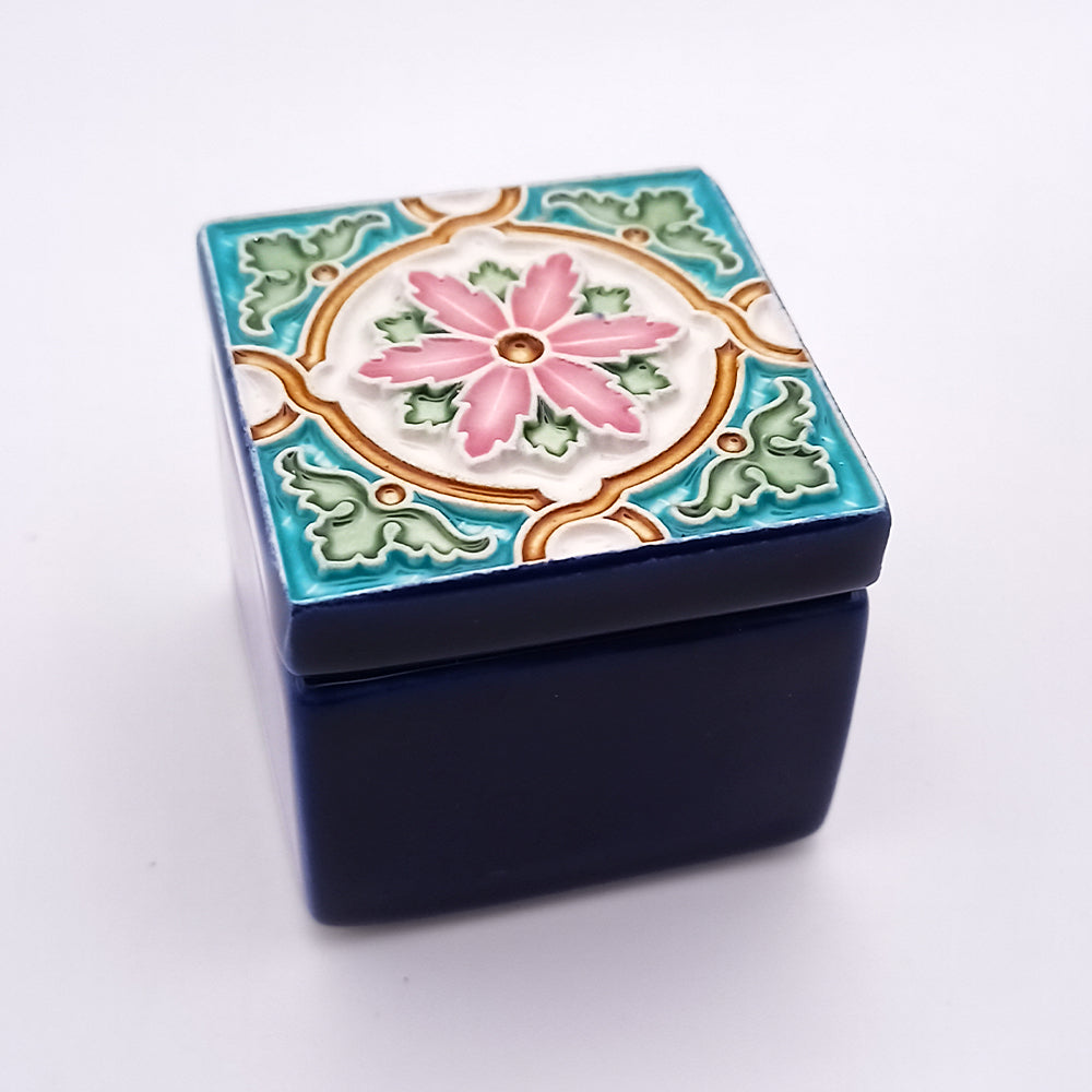 A small, square ceramic box with a deep blue base and a decorative lid featuring a floral design. The lid has a central pink flower with green leaves and gold-colored accents, framed by a blue and white geometric pattern. This Bairro Alto Ceramic Box from Tejo Shop showcases refined craftsmanship throughout its exquisite and exclusive detailing.