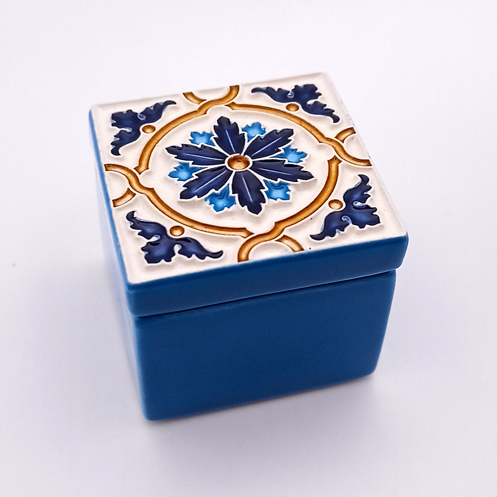 A small, square, blue ceramic box with a detachable lid. The Belém Alto Ceramic Box by Tejo Shop features an intricate, symmetrical design with dark blue and gold floral and geometric patterns against a white background. This hand-crafted piece showcases an ornate and decorative style.