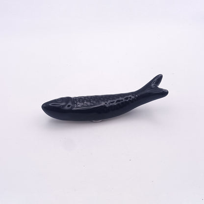 The Ceramic Sardine Fridge Magnet from Tejo Shop, reminiscent of a Portuguese icon, is shown against a plain white background. This black ceramic sardine features a textured surface replicating scales, with a clearly defined tail and minimal facial features, giving it a simple yet stylized appearance ideal for kitchen decor.