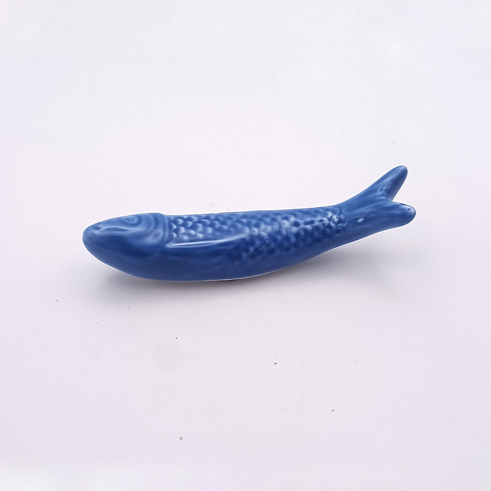 A small blue ceramic fridge magnet shaped like a sardine, lying on a plain white background. The sardine features textured scales, fins, and a tail, resembling a traditional Japanese design. Its charming simplicity makes it an ideal piece for kitchen decor. Crafted by Tejo Shop, this Ceramic Sardine Fridge Magnet embodies both functionality and aesthetic appeal.