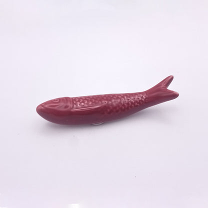 Displayed on a plain white background is a small, burgundy-colored Ceramic Sardine Fridge Magnet from Tejo Shop. This Portuguese icon features detailed scales and fins, a streamlined body with a slightly curved tail and mouth, making it a lifelike addition to your kitchen decor.