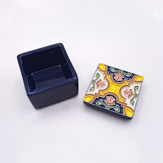 The handmade Braga Ceramic Box by Tejo Shop is a small, square, dark blue ceramic box that is open, revealing the separate lid and base. The elegant lid features a colorful, intricate pattern with yellow, green, and pink designs. The base of the box is plain and deep blue. Both pieces rest on a white background.