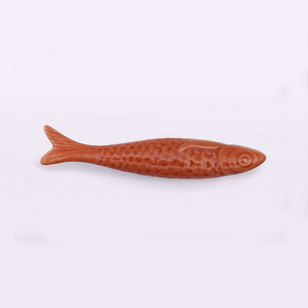 A small, brown fish-shaped object made of a textured, glossy material. It has a detailed scale pattern and features a smooth tail. Reflecting traditional craftsmanship, this piece of **Portuguese Ceramic Sardines by Tejo Shop** can be an elegant addition to your home decor against its plain white backdrop.