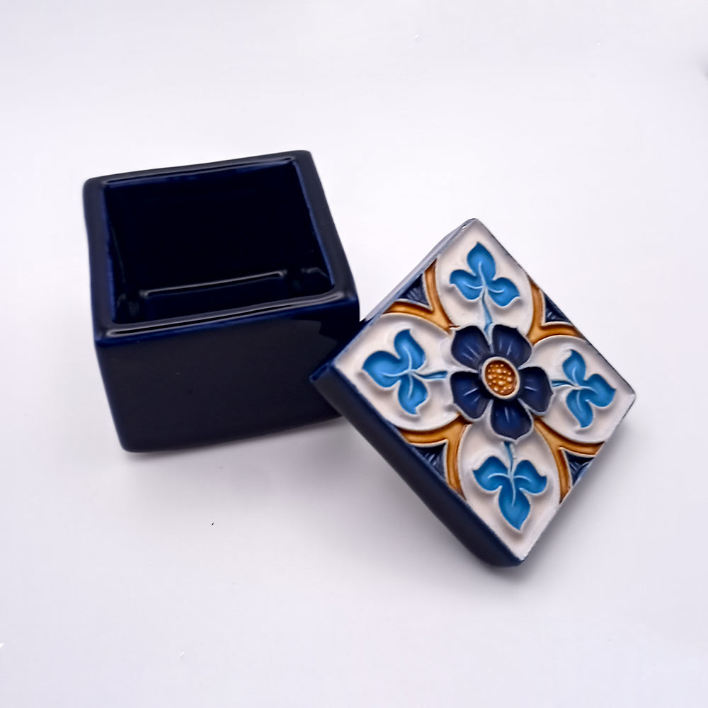 The Cascais Ceramic Box by Tejo Shop is a small, square, navy blue ceramic box exuding sophisticated elegance with its intricate floral design on the lid. The opulent past is reflected in the central blue flower surrounded by blue and gold accents on a white background. The lid is slightly lifted, revealing the empty interior.