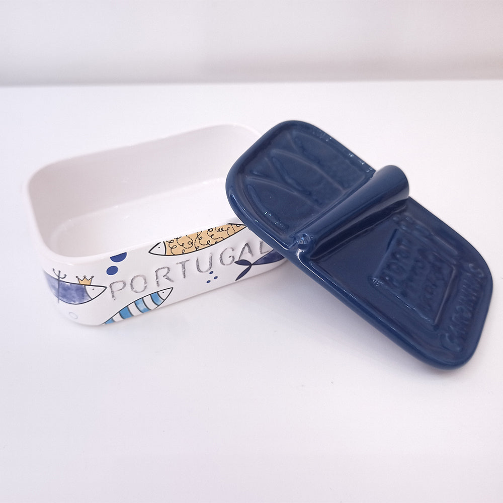 The Ceramic Soap Box - Sardine Special Edition by 100ml, designed in a rectangular shape to resemble a sardine can, is placed on a white surface. A true Portuguese-style piece, its cover is blue while the body features blue fish and the word "PORTUGAL" in blue letters. The cover is slightly open, revealing the empty white interior.