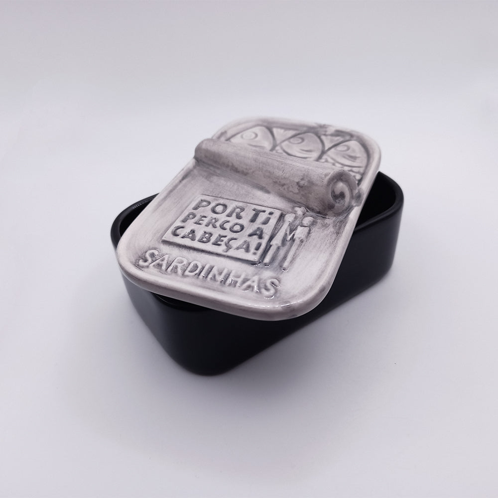 The "Aguada Edition" hand-painted ceramic soap box by 100ml features a black container with a lid resembling an open sardine can. It's embossed with scrolls and the text "POR TI PERCOLA CABEÇA - SARDINHAS," and is part of the "I'm Crazy About You" collection. The interior is empty, ready for use.