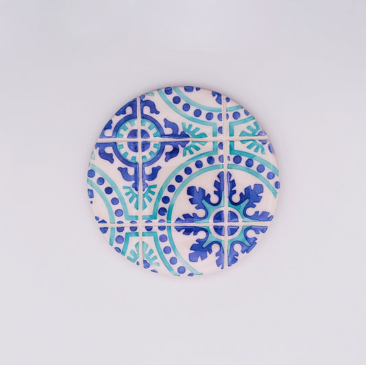 A circular ceramic coaster with a blue and turquoise pattern featuring ornate, symmetrical designs, this Chaves Ceramic Cup Pad by Tejo Shop resembles traditional Spanish or Moroccan tiles. Intricate curves and floral elements set against a white background make it a beautiful addition to any home decor.