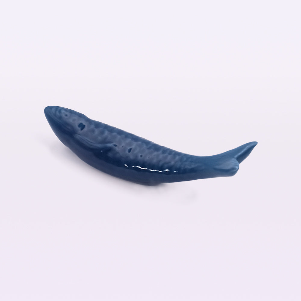 A small, dark blue **Portuguese Ceramic Sardine** lies on a light surface. The fish, inspired by traditional Portuguese ceramic sardines from **Tejo Shop**, is streamlined with a slightly raised texture on its body, giving the appearance of scales. The tail fin is pointed, and the overall shape is sleek and minimalistic—a perfect piece for home decor.