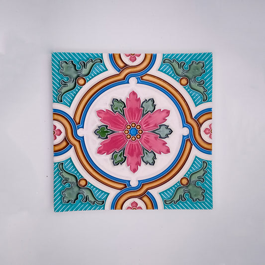 A Tejo Shop decorative backsplash tile featuring a colorful symmetrical floral design with a central pink flower surrounded by hand-painted green, blue, and orange patterns on a white background.