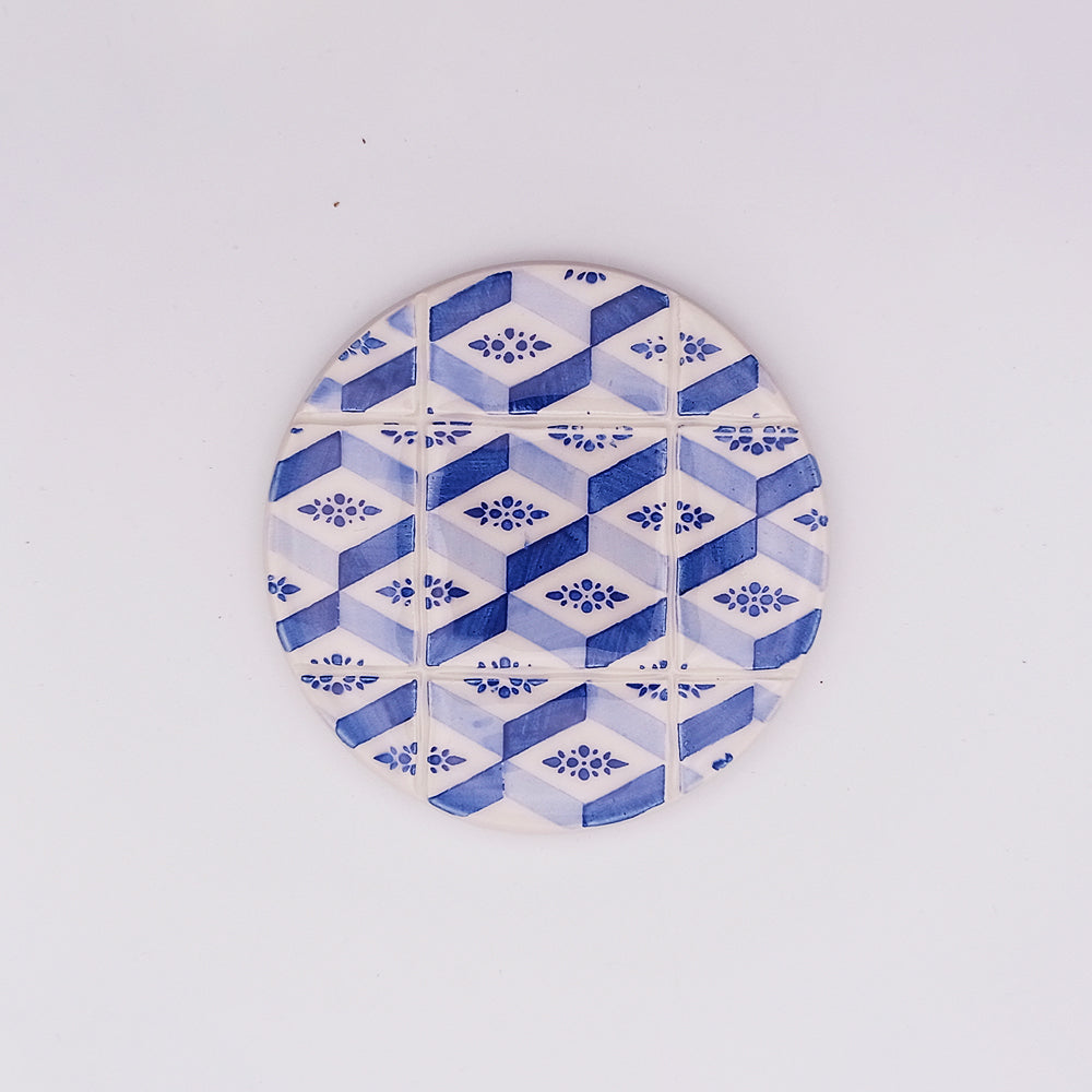 A round ceramic coaster with a geometric pattern of blue and white hexagons, each containing a small floral design at the center, arranged in a grid formation against a white background, exudes an artisanal charm, reminiscent of the hand-made Estoril Ceramic Tile Cup Pad by Tejo Shop.