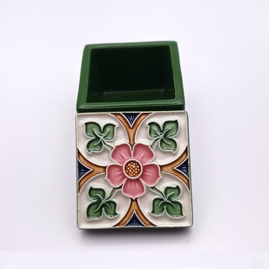 A small square Evora Ceramic Box by Tejo Shop with the lid slightly open. The lid features a floral design with a pink flower in the center, surrounded by green leaves and intricate patterns in blue, white, and orange. The base of this luxurious ceramic box is solid green.