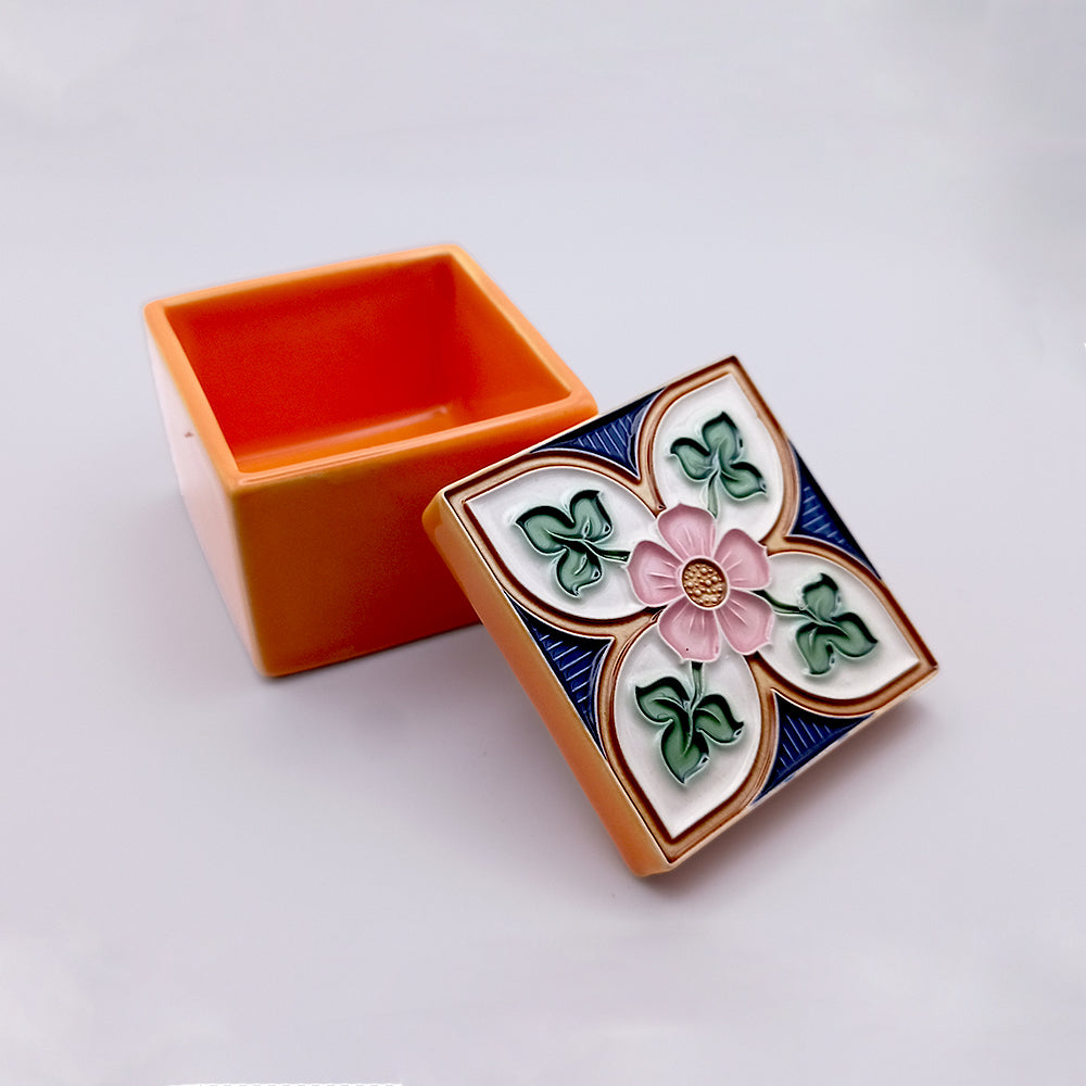 A small, square, orange Fatima Ceramic Box from Tejo Shop is shown with its lid removed and resting to the side. The handcrafted lid features a colorful, intricate floral design with a pink flower at the center surrounded by green leaves and blue accents. The background is plain white.