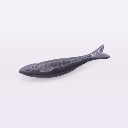 A ceramic fish-shaped dish with a glossy dark gray finish lies on a white surface. This piece of home decor is stylized to mimic the form and scales of a fish, with a raised body and detailed tail fin, reminiscent of traditional craftsmanship seen in Portuguese Ceramic Sardines by Tejo Shop.