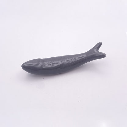 A small, smooth, and shiny gray ceramic figurine shaped like a fish, reminiscent of the Ceramic Sardine Fridge Magnet by Tejo Shop. The fish features minimal detailing and is set against a plain white background, making it ideal for kitchen decor.