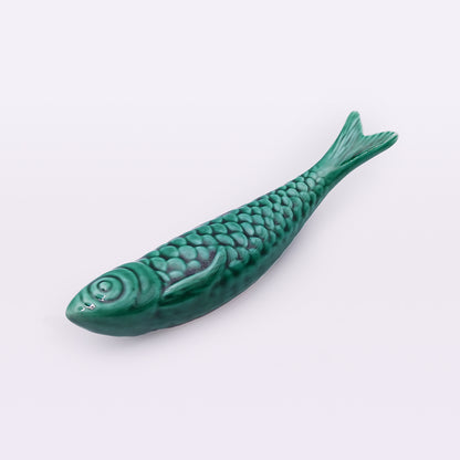 A green fish-shaped ceramic figurine lies on a plain white background. Handcrafted with detailed scales and fins, this piece showcases the traditional craftsmanship of Portuguese Ceramic Sardines by Tejo Shop. Its slightly curved body and glossy finish give it a shiny appearance.