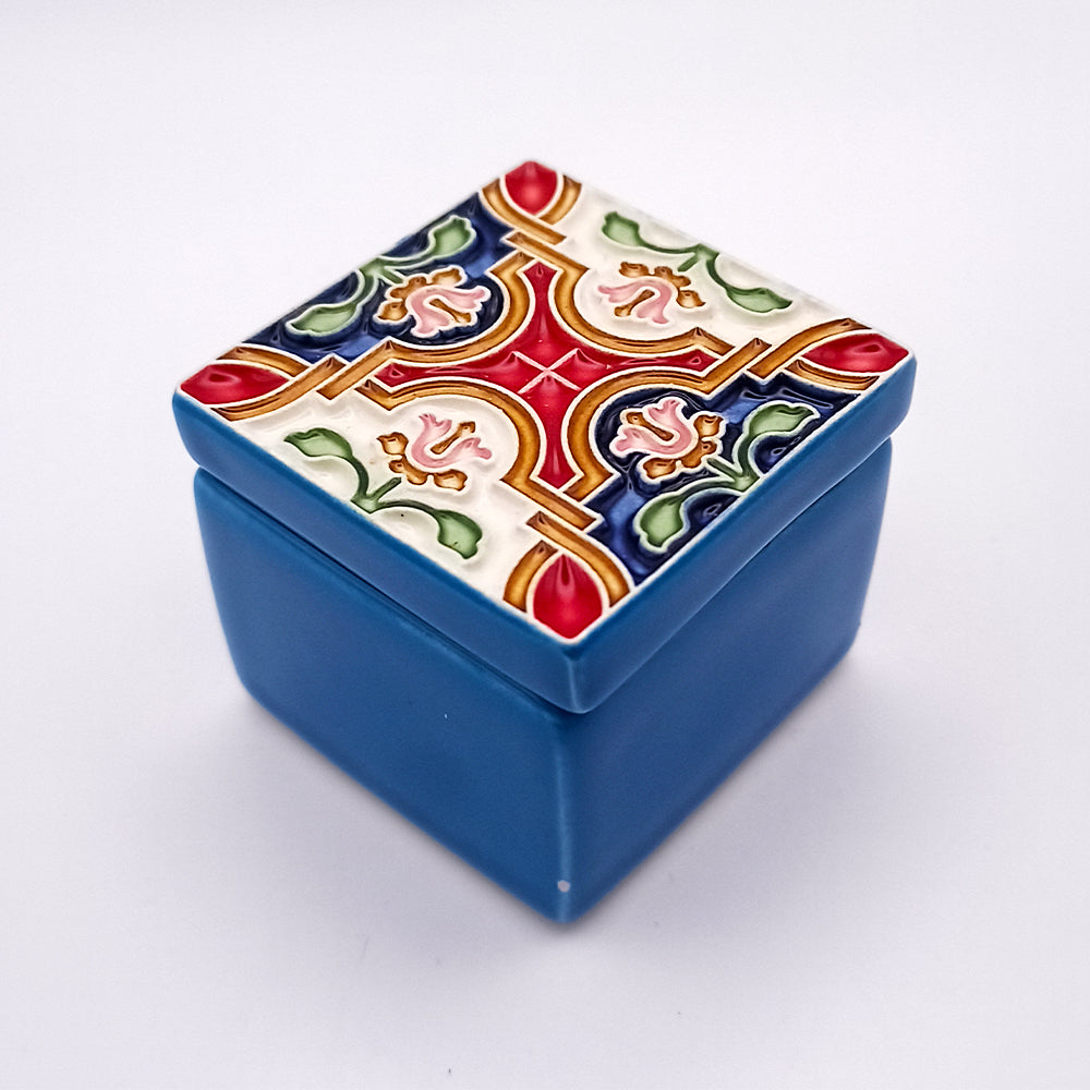 A charming ceramic box with a plain blue body and a decorative lid featuring a colorful, intricate pattern. The pattern includes red, blue, green, and yellow designs resembling a traditional, ornate motif. This small square Guimarães Ceramic Box by Tejo Shop holds significant cultural importance.