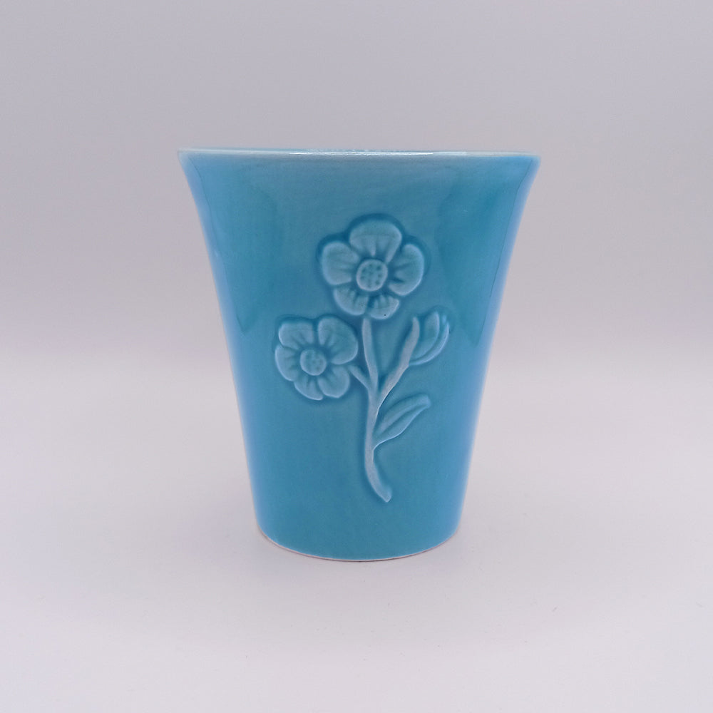 The Ceramic Mug by Tejo Shop is a small blue ceramic cup with a flared shape, featuring a unique embossed design of two flowers with stems and leaves on one side. Inspired by Portuguese art, the mug has a plain white surface background.