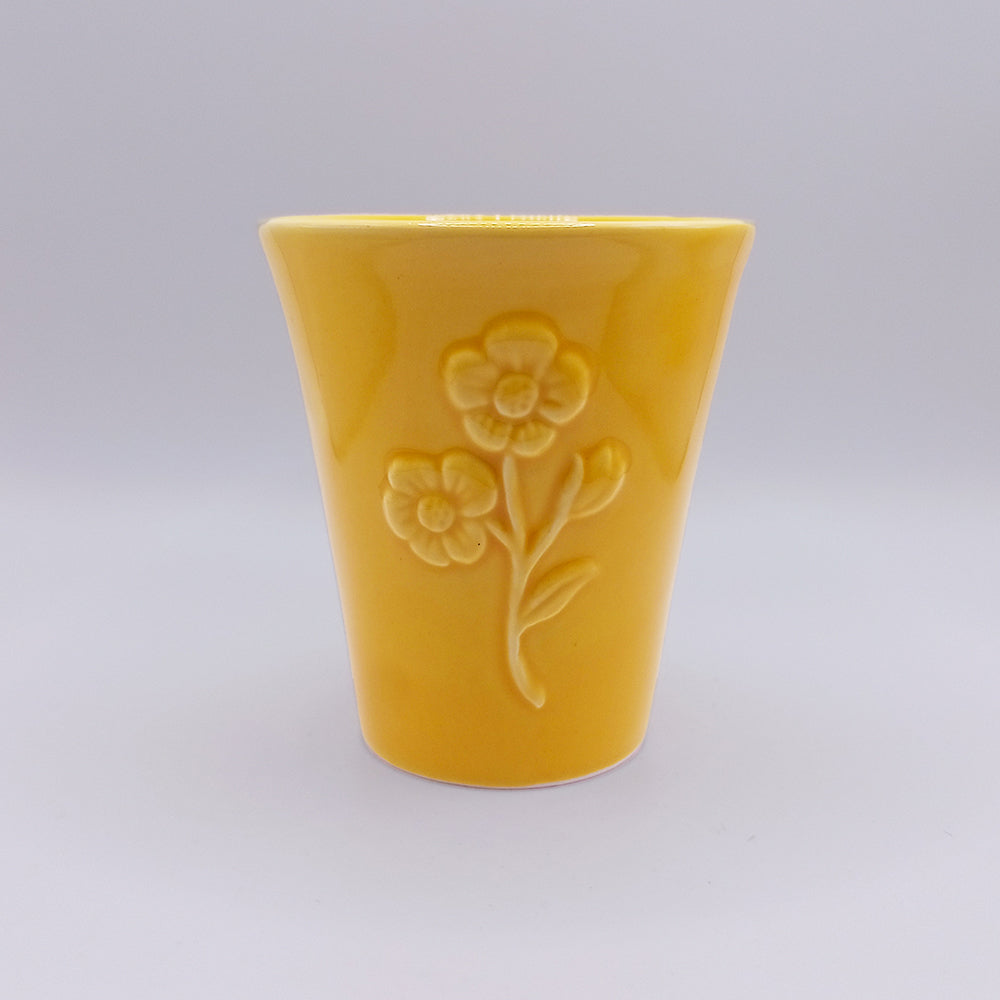 Product Description: This handmade ceramic mug from Tejo Shop features a striking yellow color with a matte finish. The front showcases an intricate floral relief design depicting two flowers and a stem, drawing inspiration from Portuguese art. It stands out beautifully against a plain, light gray gradient background.