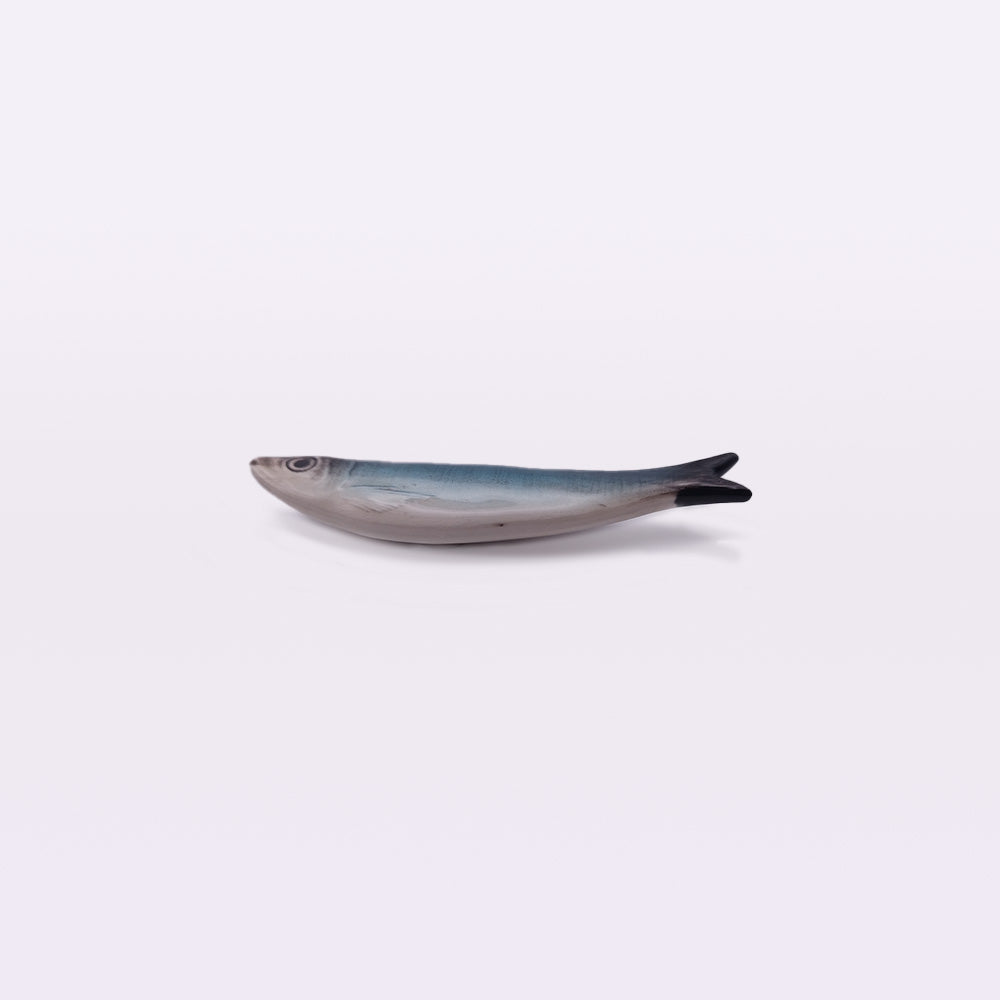 A small, lifelike Ceramic Sardine Fridge Magnet with a greyish-blue body and a dark-tipped tail is centered against a plain white background. Hand made by artisans at Tejo Shop, the fish is horizontal, facing left.