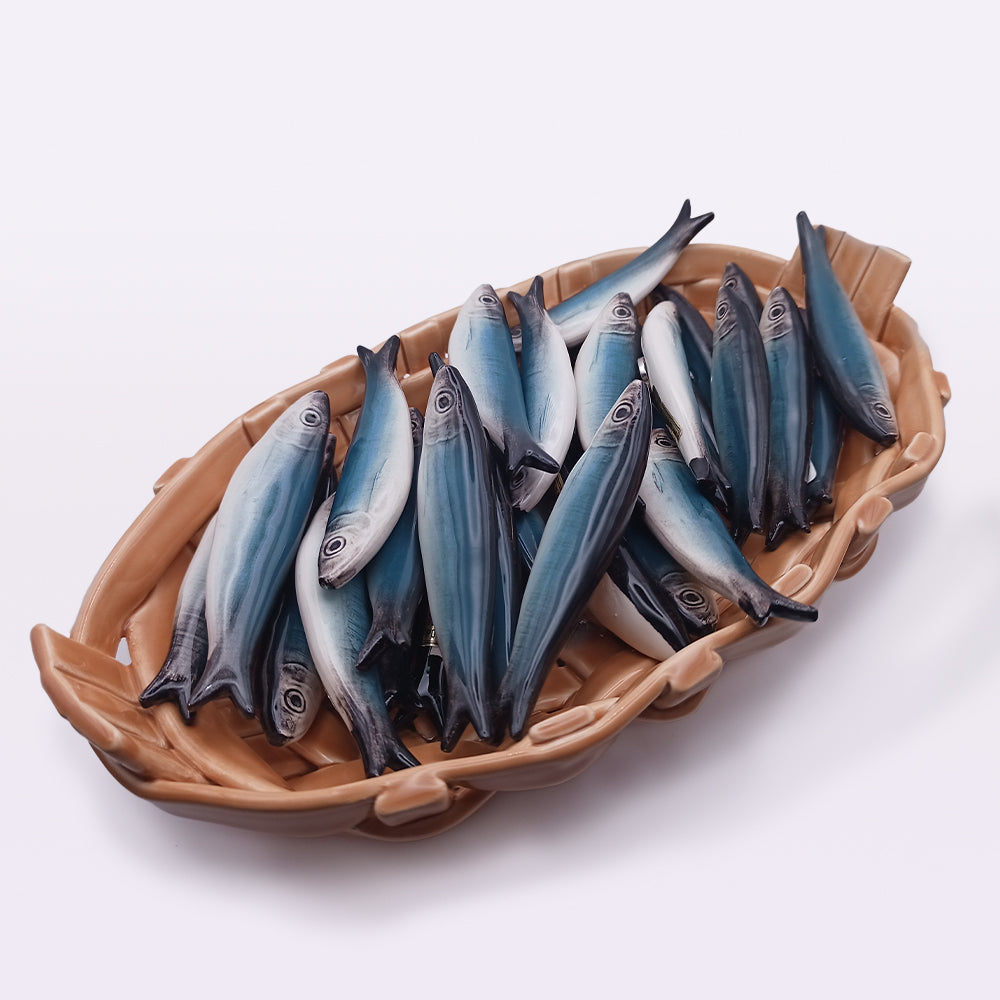 A woven basket is filled with numerous small, realistic-looking artificial fish, reminiscent of detailed Ceramic Sardine Fridge Magnets by Tejo Shop. They feature a gradient of dark to light blue colors and intricate markings, resting on a white surface.