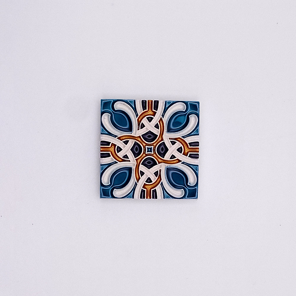 A hand-painted small ceramic tile featuring a symmetric pattern in shades of blue, white, and burnt orange on a light gray background from Tejo Shop.