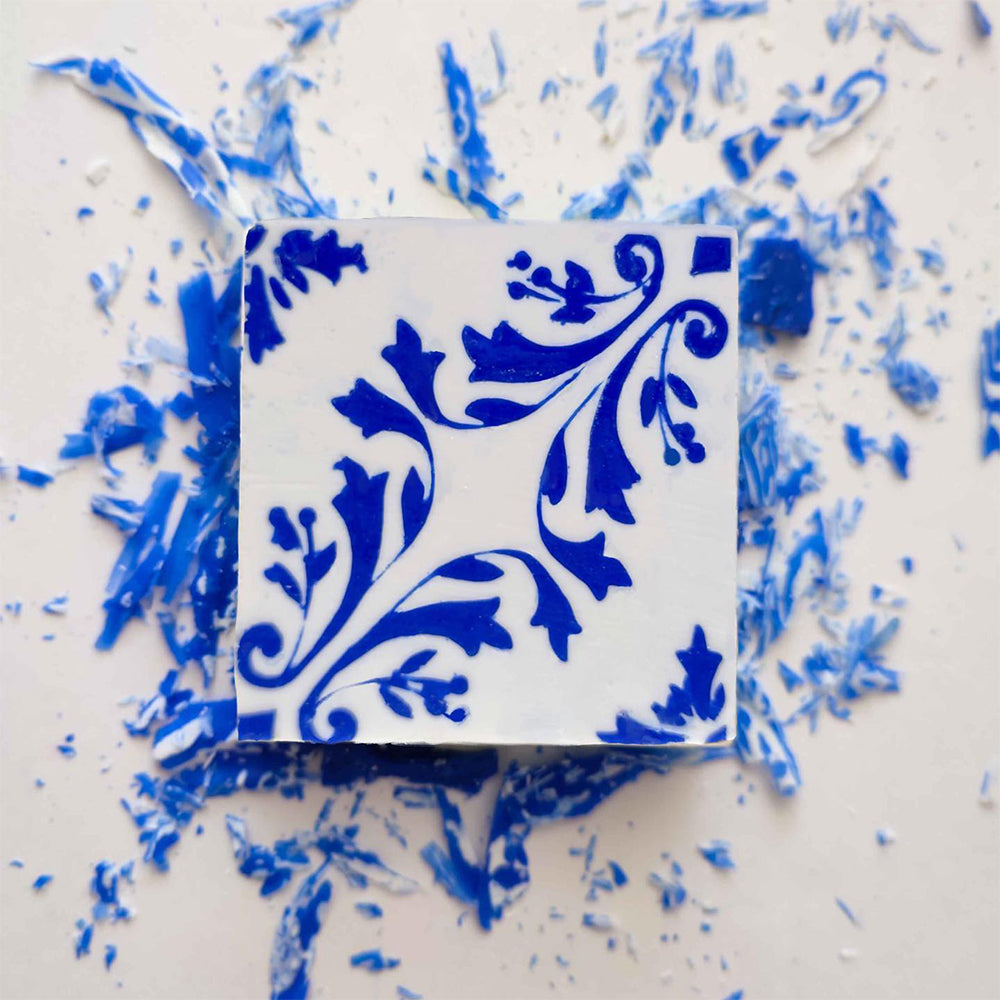 The Lisbão Amêndoa Royale Tile Soap Bar, a square soap featuring an intricate blue floral pattern, rests on a surface sprinkled with blue and white soap shavings.