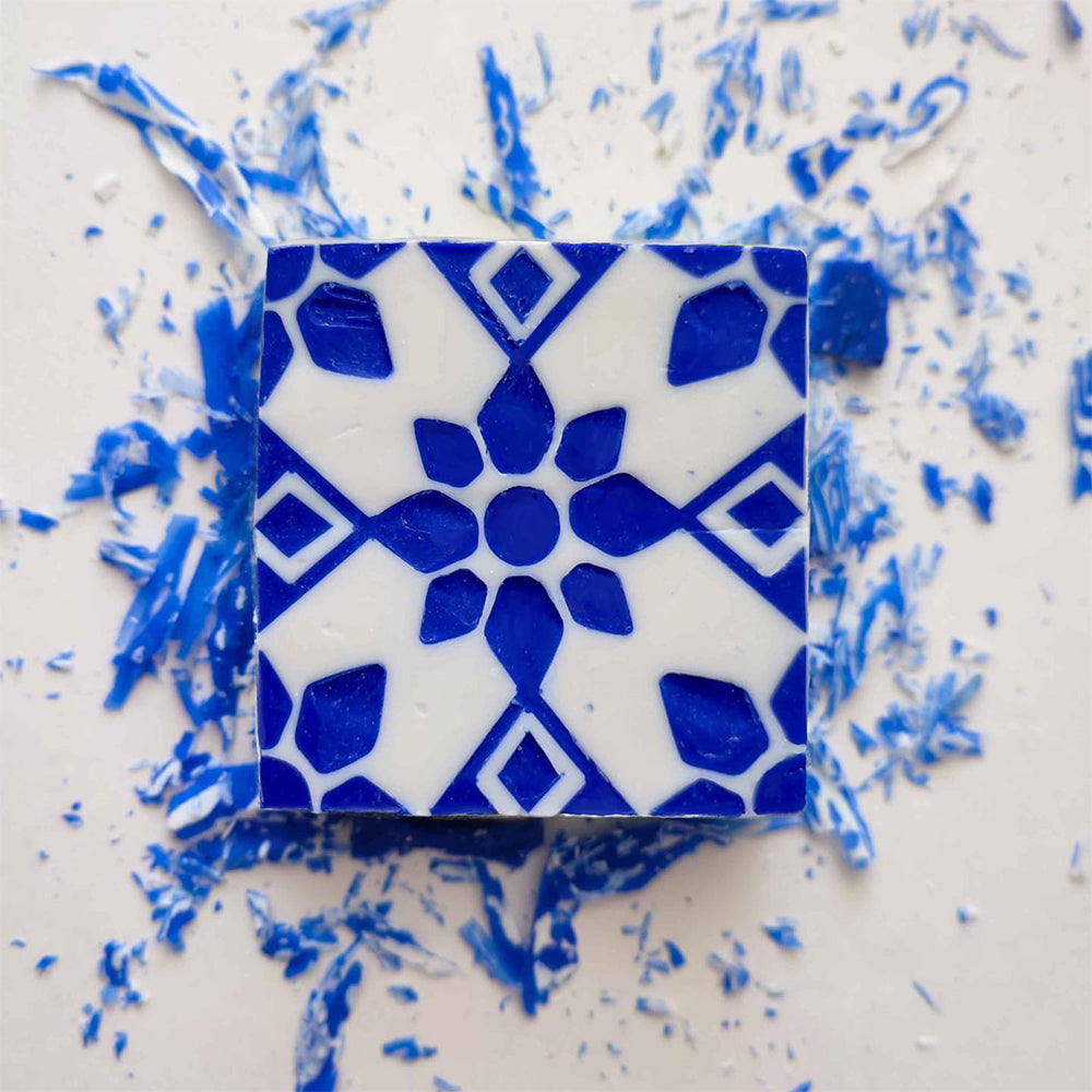 The Amêndoa Royale Tile Soap Bar by Lisbão features a square, blue and white geometric floral design, displayed with blue shavings on a white surface.
