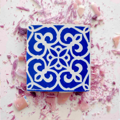 Rosa de Azulejo by Lisbão is a blue and white decorative tile soap featuring an intricate floral pattern, set among pink and purple shards and dust, capturing the charm of handmade Portuguese tiles.