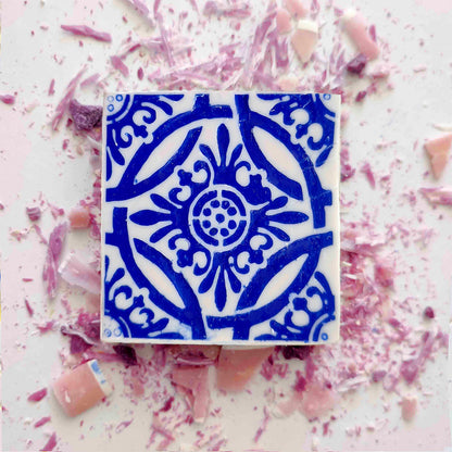 A square Lisbão Rosa de Azulejo handmade Portuguese tile soap, featuring an intricate blue pattern, rests on a white surface amid scattered pink and purple candy shavings and small broken pieces.