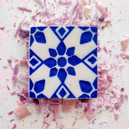 Rosa de Azulejo by Lisbão: a square white artisan soap inspired by bold blue geometric floral Portuguese tiles, is centered on a light surface, with pink and purple shards scattered around.
