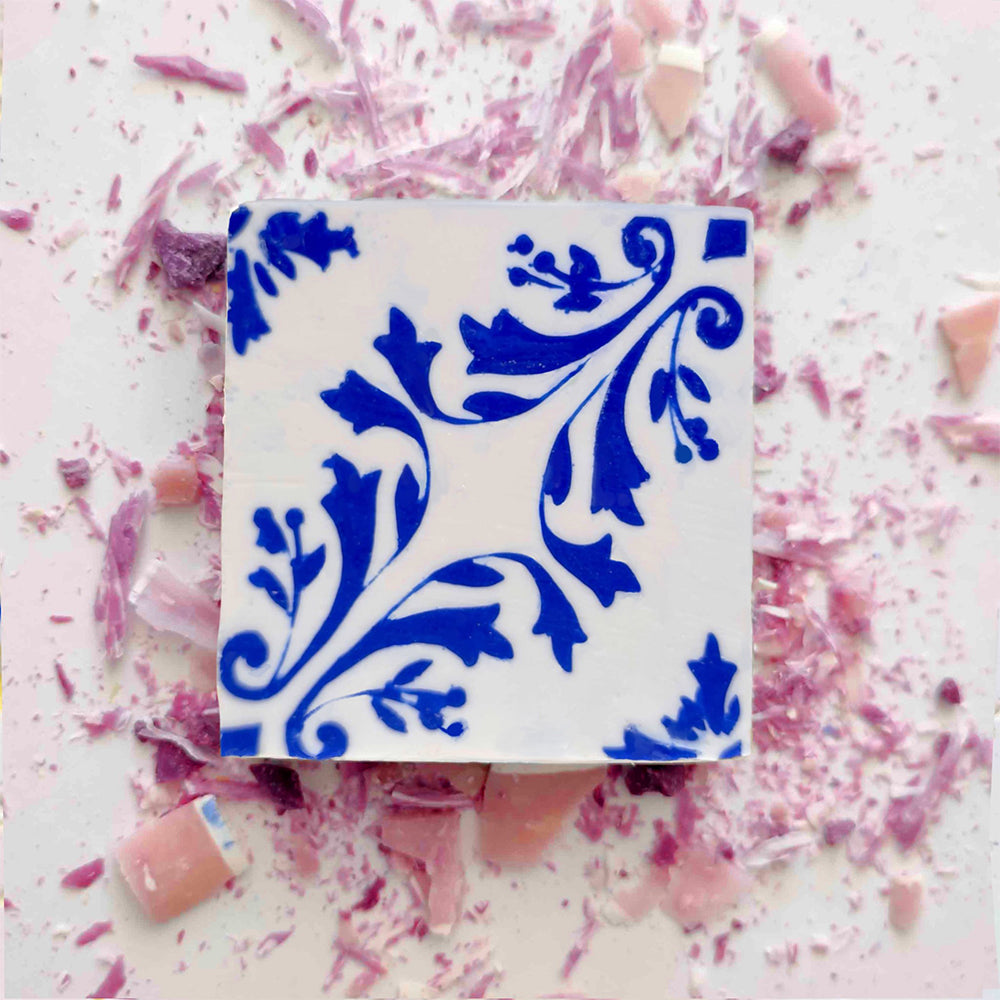 Rosa de Azulejo by Lisbão is a handmade square soap inspired by Portuguese tiles, featuring white and blue floral patterns and presented amid pink and purple fragments, capturing the charm of artisanal craftsmanship.